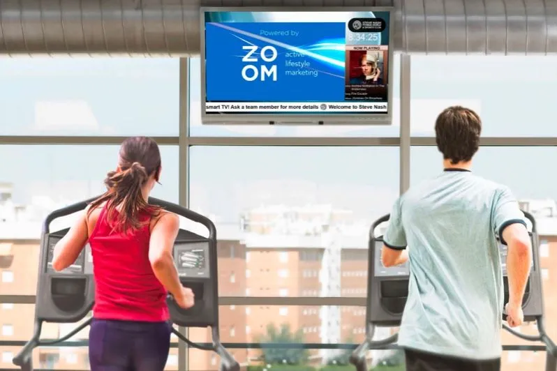 Socios de un gimnasio en máquinas de cardio bajo una pantalla en la pared